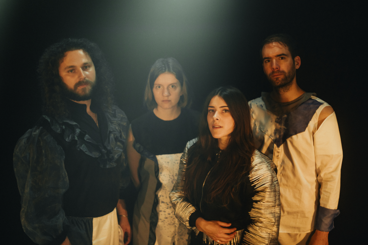 die vier Musiker:innen von BARANY stehen vor einem schwarzen Hintergrund in einem orange/blauen Licht und schauen in die Kamera