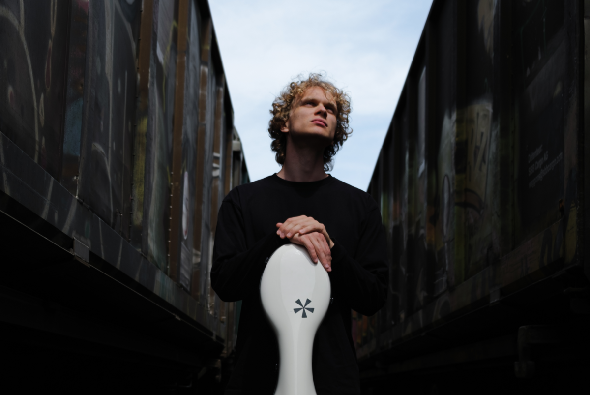 Maksim Barbash sthet zwischen zwei dunklen Wänden, über ihm strahlt der blaue Himmel, er stützt seine Hände auf seinem weissen Cello Koffer ab und schaut verträumt nach oben.