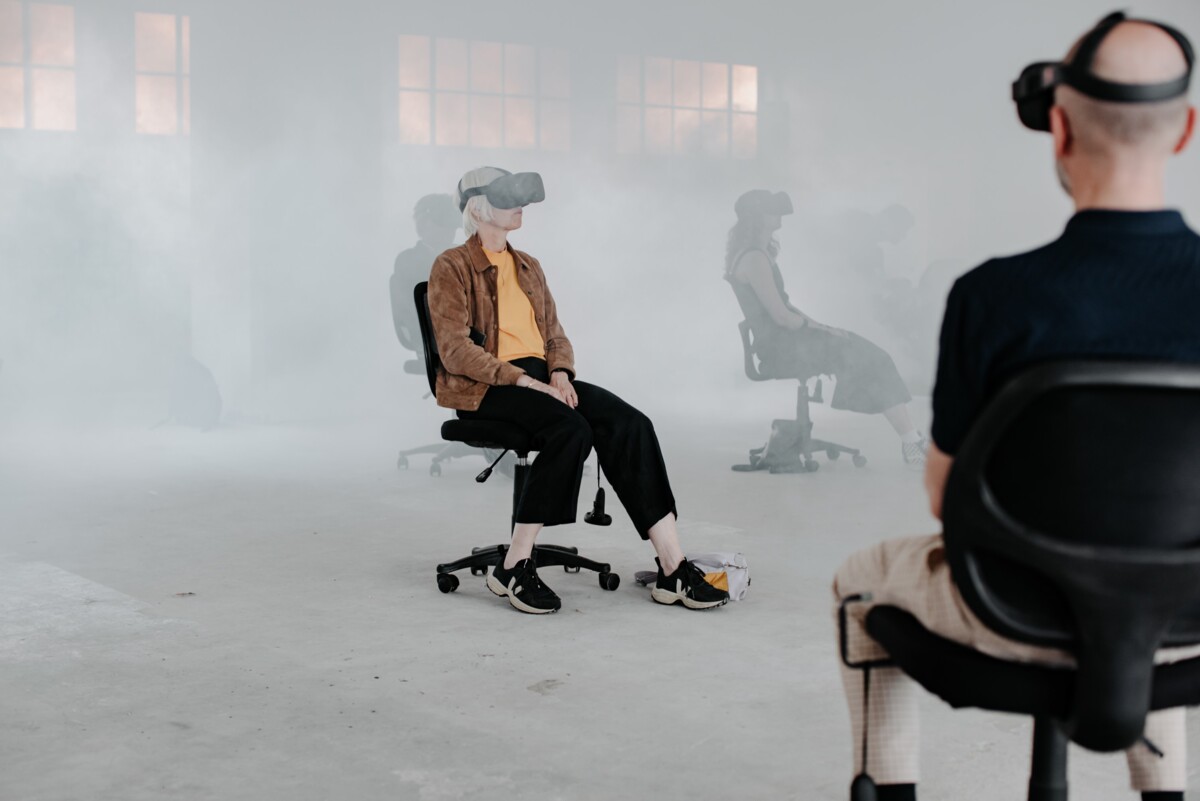 Bild der Performance Ascension. Zwei Personen mit VR Brillen sitzen auf Drehtstühlen in einem weissen Raum mit Nebel.