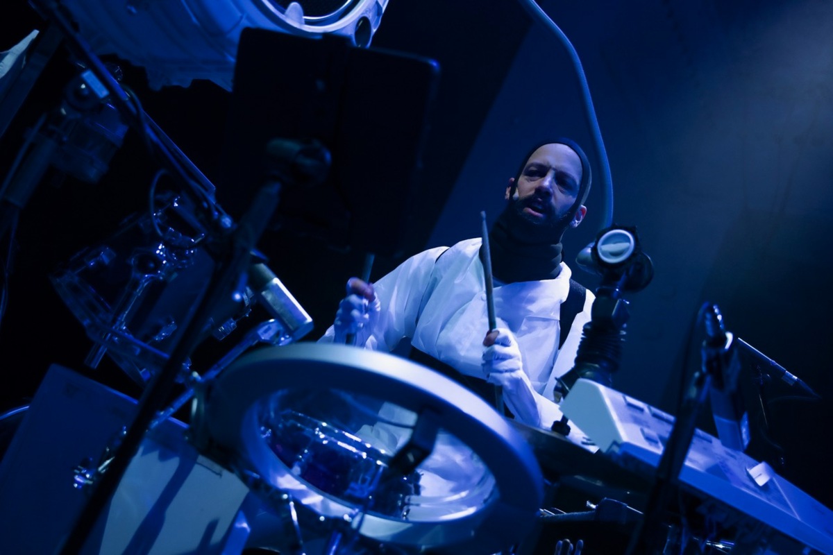 Julien Mégroz spielt Schlagzeug in blauem Bühnenlicht und weissem Anzug